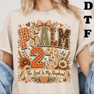 May include: A beige t-shirt featuring a graphic design with the text PSALM 23 and The Lord Is My Shepherd. The design includes a plaid bow, autumn-colored floral patterns, wheat stalks, and hearts in shades of orange, brown, green, and cream.