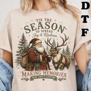 May include: A beige t-shirt featuring a graphic of Santa Claus and a reindeer in a forest. The text reads 'TIS THE SEASON TO SPREAD Joy & Kindness MAKING MEMORIES THAT LAST A LIFETIME'. Santa wears a red coat and carries a sack near pine trees.
