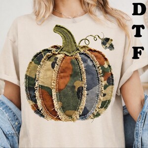 Peut inclure: Un t-shirt beige arborant un grand motif de citrouille avec un imprimé camouflage et des bords en tissu effiloché. La citrouille possède une tige verte et une vrille. Les lettres DTF sont imprimées en noir sur le côté supérieur droit.