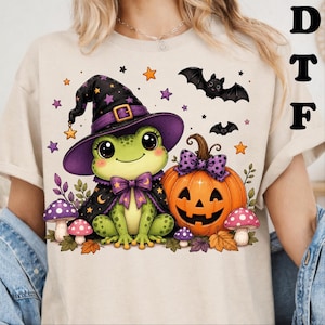 May include: A beige t-shirt featuring a graphic of a green frog wearing a purple witch hat and cape with a bow tie. Next to the frog is an orange jack-o'-lantern with a purple bow, surrounded by small mushrooms, autumn leaves, stars, and bats.