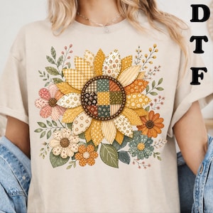 May include: A beige short-sleeve t-shirt featuring a large central graphic of a sunflower with a patchwork-style center and petals in various yellow, orange, and green patterns, surrounded by smaller flowers and leafy stems in muted tones.