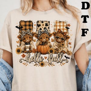 May include: A beige t-shirt featuring a graphic of three Highland cows wearing bows, sweaters, and accessories. The design includes pumpkins, sunflowers, autumn leaves, bats, and the text "hello fall" in a cursive font, set against plaid backgrounds.
