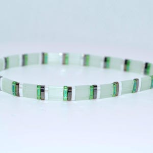May include: A beaded bracelet featuring a repeating pattern of rectangular pale mint green glass beads, thin white spacers, and small metallic dark grey and vibrant green accent beads arranged in a continuous circular band design.