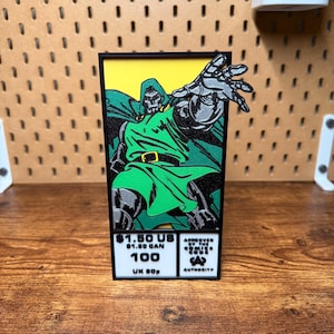 May include: A 3D-printed rectangular display featuring Doctor Doom in a green tunic and silver armor against a yellow background. The base includes text: $1.50 US, $1.80 CAN, 100, UK 50p, and the Comics Code Authority seal in black and white.