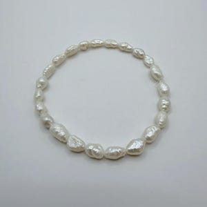 Könnte beinhalten: Ein kreisförmiges Armband, das aus zahlreichen weißen, unregelmäßig geformten Barockperlen besteht. Jede Perle weist eine ausgeprägte, organische Textur mit einer glänzenden, reflektierenden Oberfläche auf, die zu einer durchgehenden, flexiblen Schlaufe aufgereiht sind.
