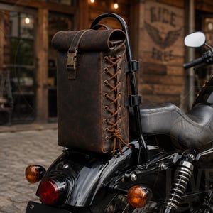 May include: A dark brown leather roll-top motorcycle sissy bar bag featuring a vertical lace-up design with metal eyelets on the side and a brass-toned buckle closure on the front strap, securely mounted to the back of a black motorcycle seat.