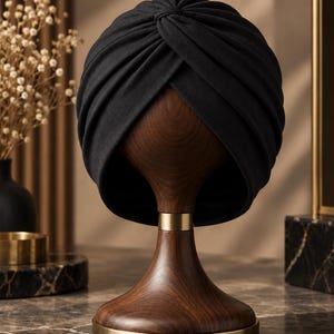 May include: A solid black turban hat featuring a gathered, knotted front design. The head covering is displayed on a dark brown wooden mannequin head stand with a smooth, polished finish and a metallic gold base accent at the neck and bottom.
