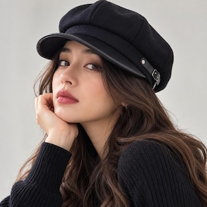 Puede incluir: Una gorra de estilo marinero en color negro con una corona estructurada y una visera rígida. El accesorio presenta una tira decorativa negra en la parte frontal, asegurada con una pequeña hebilla metálica plateada y un remache a juego.