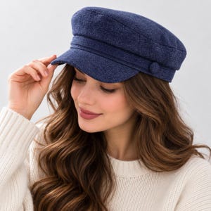 May include: A navy blue newsboy cap featuring a textured fabric finish, a structured brim, and a decorative band across the front with a matching circular button detail on the side. The cap has a rounded crown and a classic flat-top silhouette.