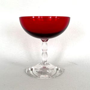 May include: A vintage red glass coupe. The bowl is a deep red, transitioning to a darker shade at the base. It has a clear, ornate stem with a twisted design, and a circular base. The coupe is suitable for serving champagne.