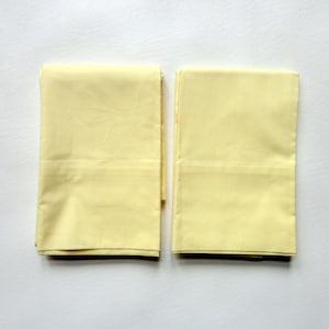 May include: Two folded, pale yellow pillowcases are displayed on a white surface. The pillowcases are rectangular and appear to be made of a smooth fabric. The folded edges are visible, and the overall presentation is neat and clean.