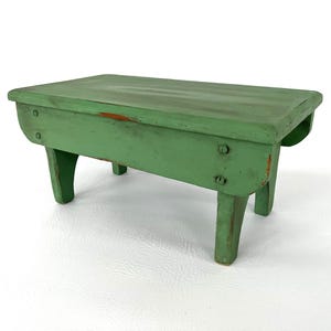 Puede incluir: Un pequeño taburete de madera verde rectangular. El taburete tiene cuatro patas curvas y una parte superior plana. La pintura verde está desgastada, mostrando rastros de la madera debajo. El taburete mide aproximadamente 46 cm de largo.