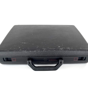 May include: A black Samsonite briefcase with a textured surface. The briefcase has a black handle and a lock. The corners have red accents. The briefcase is rectangular and sits on a white surface.