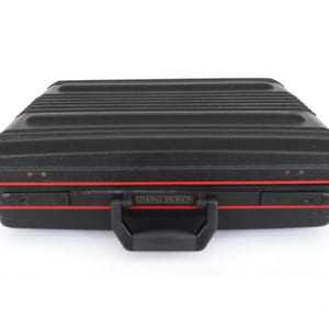 May include: Black hard-sided briefcase with a ridged top and red trim. The case has a black handle and a metal nameplate that reads "SEIMON". The briefcase is closed and sits on a white background.