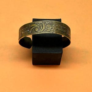 May include: A metallic cuff bracelet with a brass-toned finish featuring embossed designs and text. The engraving includes the words A CENTURY OF PROGRESS, TRAVEL AND TRANSPORT, and depictions of stylized figures and architectural structures.