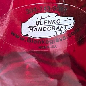 May include: A vibrant red glass object featuring an oval sticker with the text 304-743-9081, BLENKO HANDCRAFT, www.Blenkoglass.com, and MADE IN U.S.A. The sticker is white with black lettering and is applied directly to the smooth, translucent red surface.