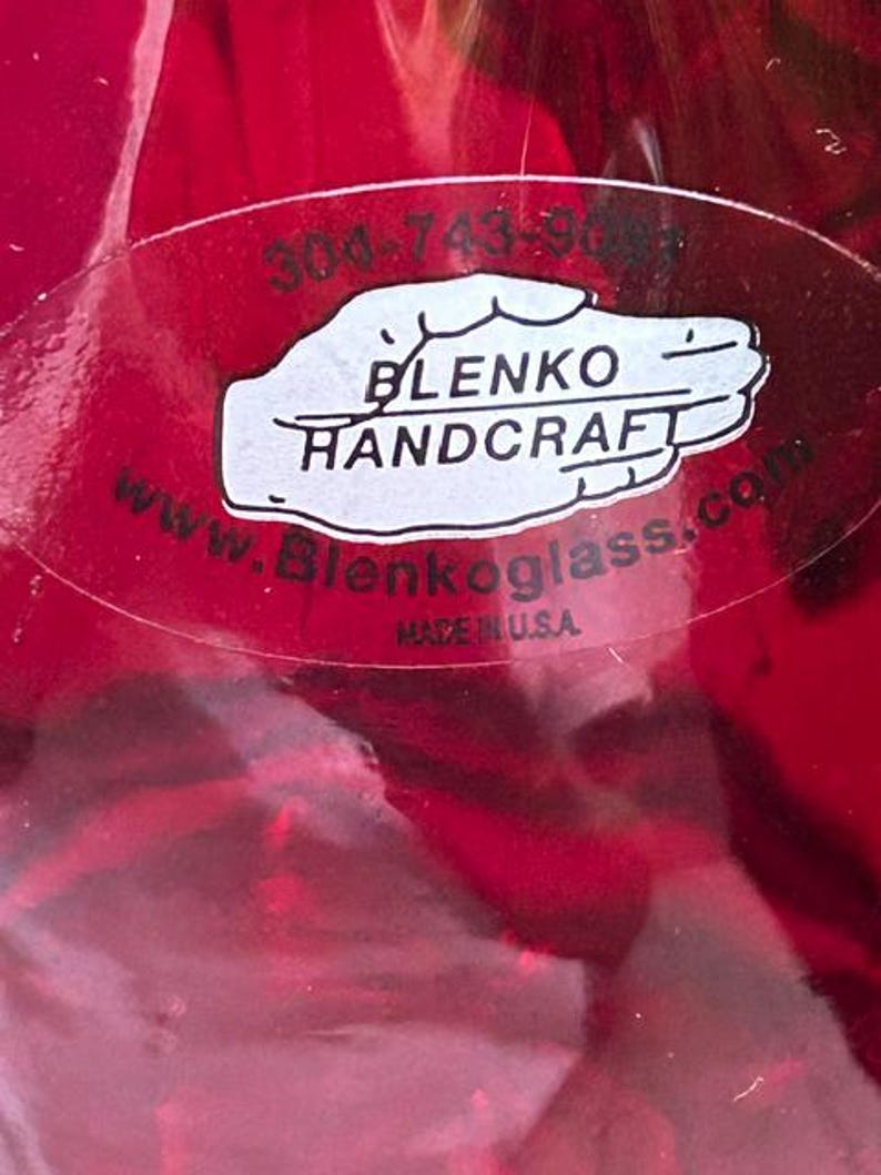 May include: A vibrant red glass object featuring an oval sticker with the text 304-743-9081, BLENKO HANDCRAFT, www.Blenkoglass.com, and MADE IN U.S.A. The sticker is white with black lettering and is applied directly to the smooth, translucent red surface.