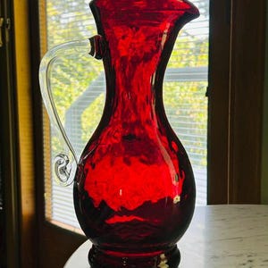 May include: A vibrant ruby red glass pitcher featuring a textured, dimpled surface and a clear, curved glass handle. The vessel has a wide, flared rim and a rounded body that tapers toward a circular base, showcasing a translucent finish.