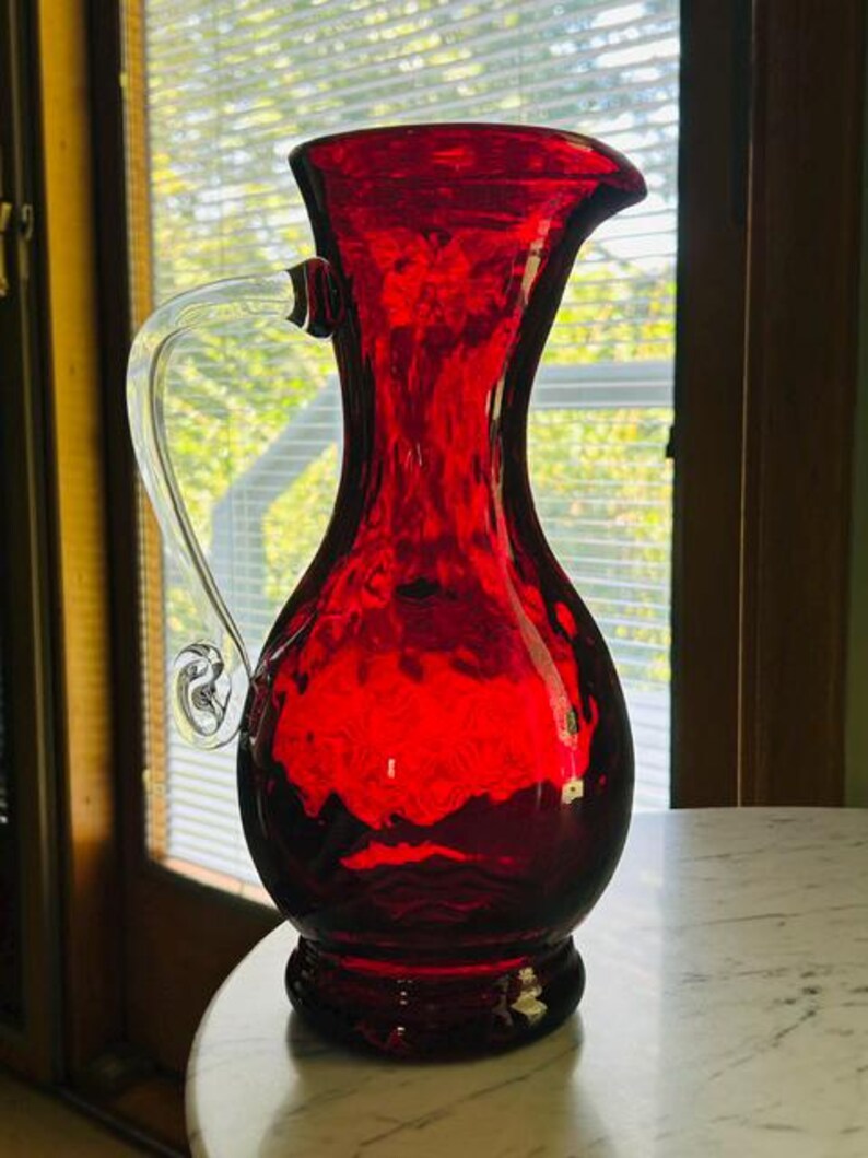 May include: A vibrant ruby red glass pitcher featuring a textured, dimpled surface and a clear, curved glass handle. The vessel has a wide, flared rim and a rounded body that tapers toward a circular base, showcasing a translucent finish.