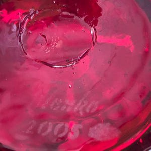 May include: A deep red, translucent glass surface featuring an etched signature that reads "Lenko" followed by the year "2005" engraved directly into the material. The glass displays a smooth, reflective texture with subtle internal bubbles.