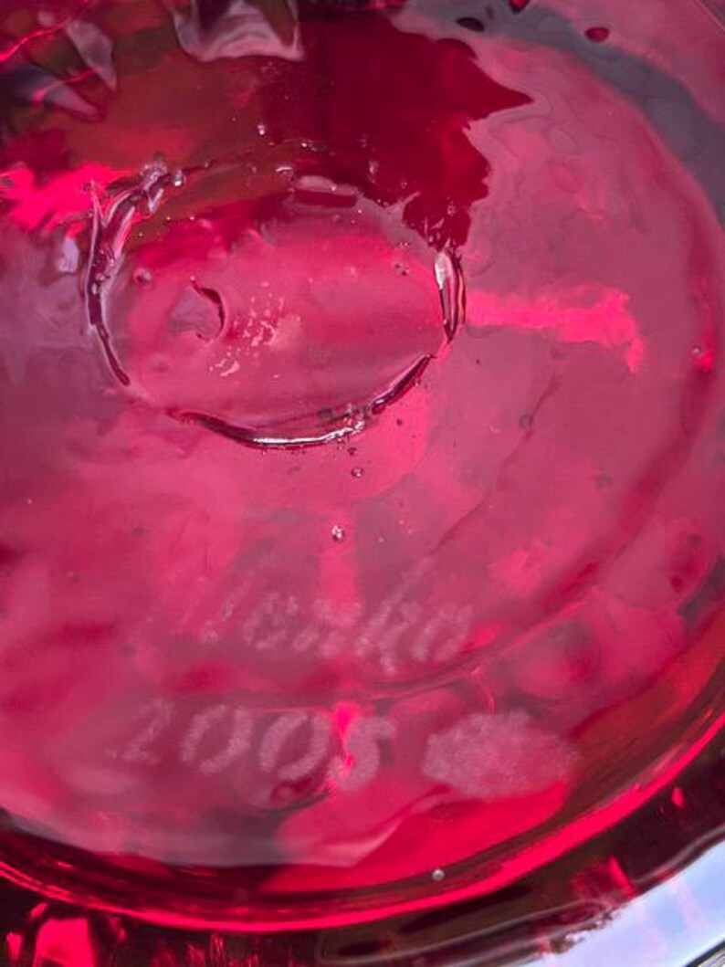May include: A deep red, translucent glass surface featuring an etched signature that reads "Lenko" followed by the year "2005" engraved directly into the material. The glass displays a smooth, reflective texture with subtle internal bubbles.