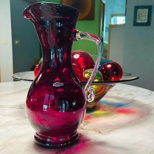 May include: A deep ruby red glass pitcher with a bulbous base, a slender neck, and a flared rim. The pitcher features a clear glass handle with a decorative swirl at the bottom and a small, rectangular label attached to the front of the body.