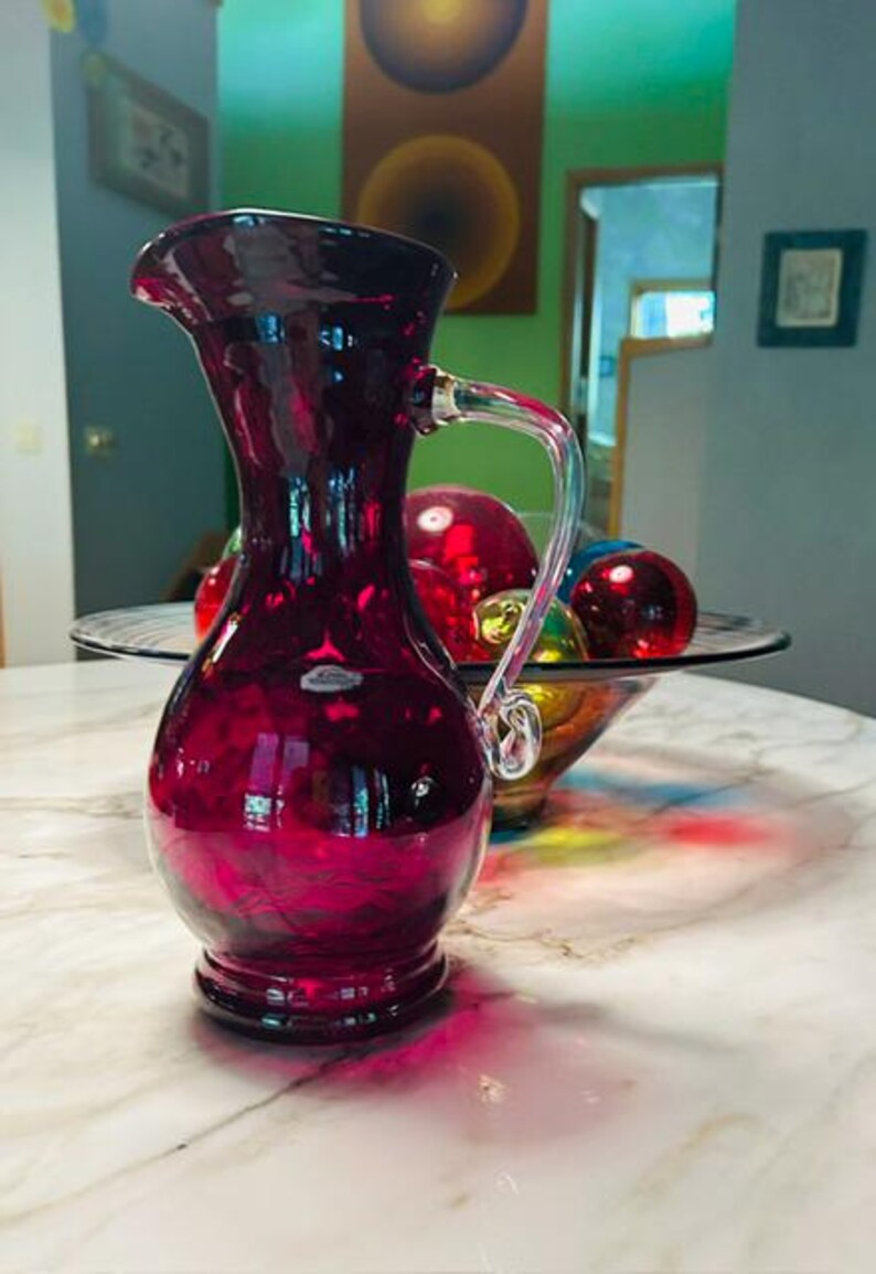May include: A deep ruby red glass pitcher with a bulbous base, a slender neck, and a flared rim. The pitcher features a clear glass handle with a decorative swirl at the bottom and a small, rectangular label attached to the front of the body.
