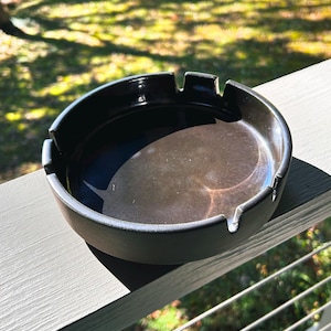 May include: A round, black ceramic ashtray featuring a smooth, glossy interior basin and a matte exterior finish. The rim is designed with three distinct notches for resting items, creating a functional and circular silhouette.