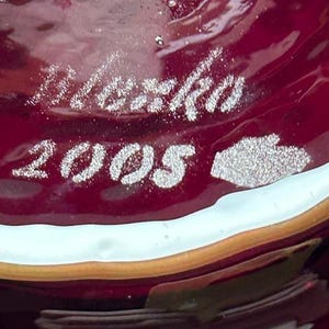 May include: A deep red glass surface featuring an etched signature that reads "Marko" above the year "2005" and a small hand-shaped icon. The glass has a smooth, glossy texture with subtle internal bubbles and a light-colored rim along the edge.