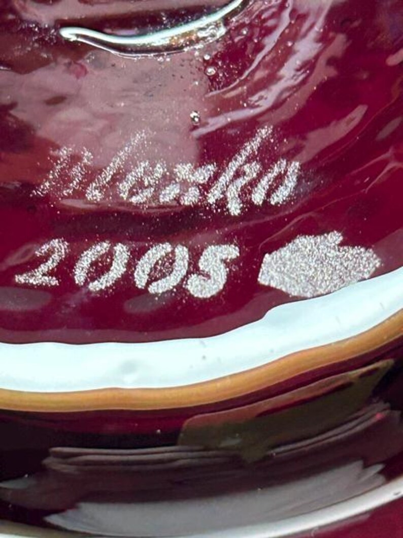 May include: A deep red glass surface featuring an etched signature that reads "Marko" above the year "2005" and a small hand-shaped icon. The glass has a smooth, glossy texture with subtle internal bubbles and a light-colored rim along the edge.