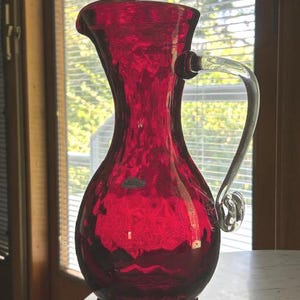 May include: A vibrant ruby red glass pitcher featuring a textured, dimpled surface and a clear, curved glass handle. The vessel has a wide, rounded base that tapers into a slender neck with a flared pouring spout and a small oval label.