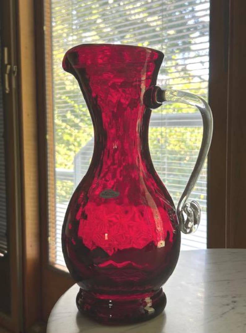 May include: A vibrant ruby red glass pitcher featuring a textured, dimpled surface and a clear, curved glass handle. The vessel has a wide, rounded base that tapers into a slender neck with a flared pouring spout and a small oval label.