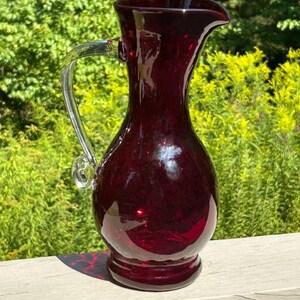 May include: A deep red glass pitcher featuring a bulbous body, a flared rim with a pouring spout, and a clear glass handle attached to the side. The pitcher has a smooth, reflective surface and a circular base, showcasing a rich, translucent crimson hue.