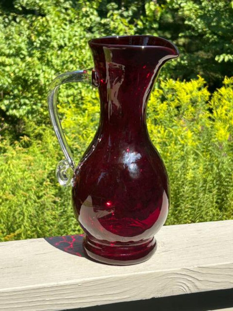May include: A deep red glass pitcher featuring a bulbous body, a flared rim with a pouring spout, and a clear glass handle attached to the side. The pitcher has a smooth, reflective surface and a circular base, showcasing a rich, translucent crimson hue.
