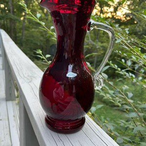 May include: A deep red glass pitcher with a bulbous body, a flared rim, and a clear glass handle. The pitcher features a small, rectangular white sticker on the front surface. The glass has a smooth, reflective finish and a pedestal base.