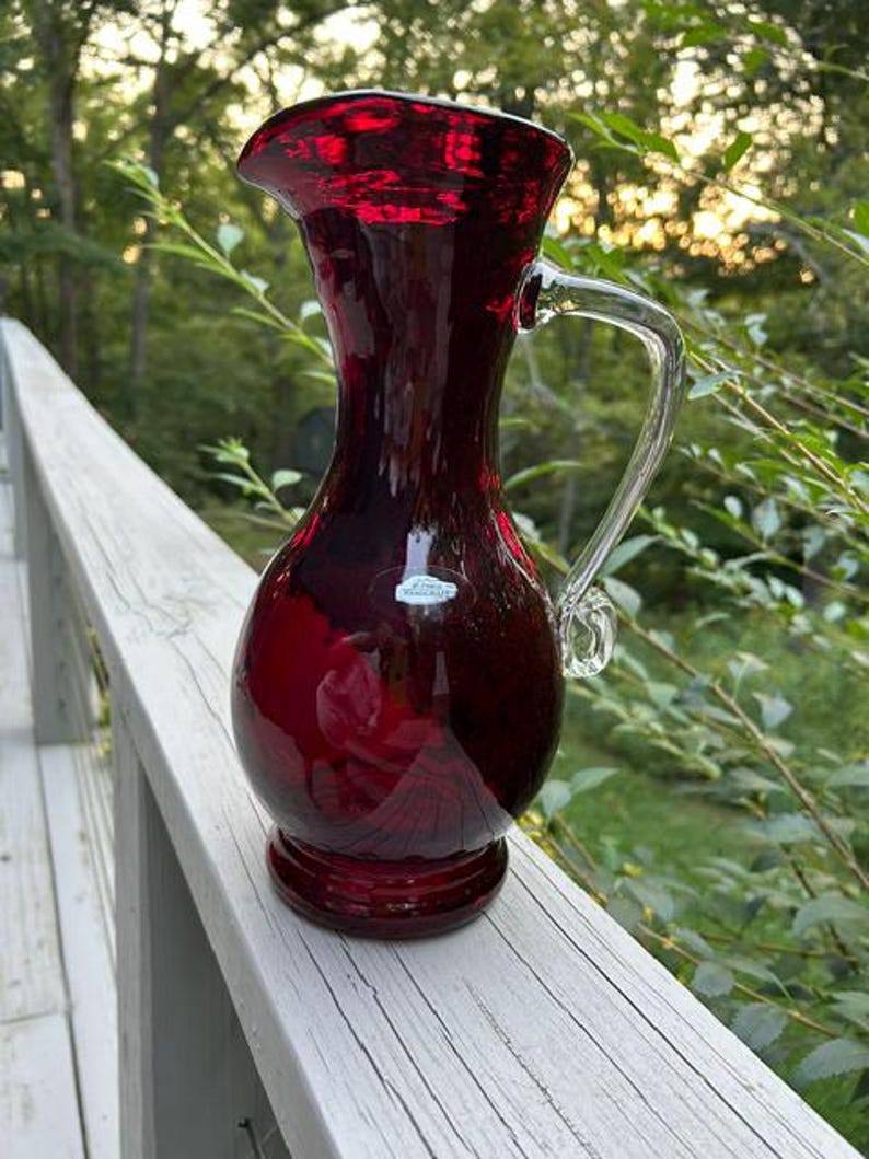 May include: A deep red glass pitcher with a bulbous body, a flared rim, and a clear glass handle. The pitcher features a small, rectangular white sticker on the front surface. The glass has a smooth, reflective finish and a pedestal base.