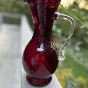 May include: A deep red glass pitcher with a textured, wavy surface, a flared rim, and a clear glass handle. The pitcher features a small, oval-shaped white sticker on the front with the text Blenko Handcraft printed in black lettering.