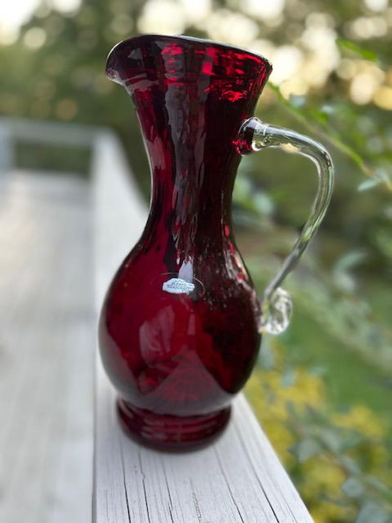 May include: A deep red glass pitcher with a textured, wavy surface, a flared rim, and a clear glass handle. The pitcher features a small, oval-shaped white sticker on the front with the text Blenko Handcraft printed in black lettering.
