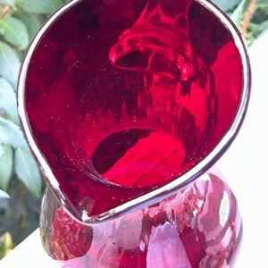 May include: A deep ruby red glass pitcher with a wide, rounded opening and a prominent pouring spout. The glass is translucent, allowing light to pass through the vibrant red color, and features a smooth, polished surface throughout the vessel.