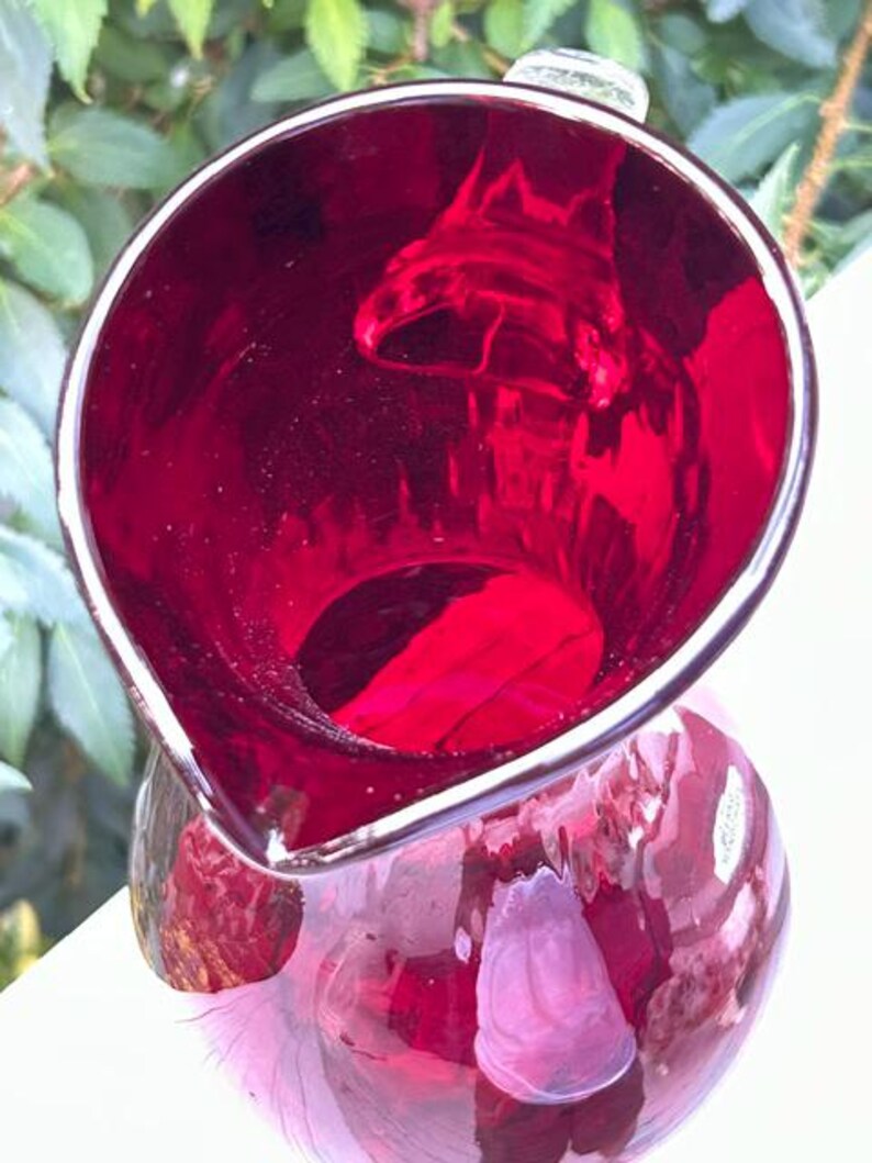 May include: A deep ruby red glass pitcher with a wide, rounded opening and a prominent pouring spout. The glass is translucent, allowing light to pass through the vibrant red color, and features a smooth, polished surface throughout the vessel.