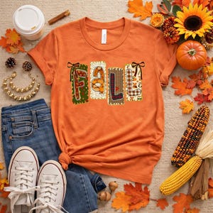 Könnte beinhalten: Ein orangefarbenes Kurzarm-T-Shirt mit dem Wort FALL in großen, dekorativen Buchstaben. Jeder Buchstabe hat ein einzigartiges Muster, darunter Kürbisse, Karomuster und Herbstblätter. Das T-Shirt hat einen Rundhalsausschnitt.