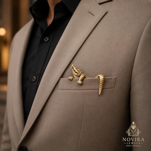 May include: A gold-toned metal crocodile brooch positioned on the breast pocket of a beige blazer worn over a black shirt. The crocodile figure features textured scales and is designed to appear as if it is emerging from the pocket opening.