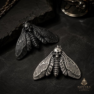 Puede incluir: Dos colgantes de polilla de metal plateado con un diseño de calavera detallado en el tórax. Cada polilla tiene alas texturizadas y veteadas con un cuerpo segmentado. En la esquina inferior derecha aparece el texto NOVIRA JEWELS con un diamante.