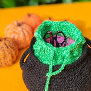 May include: A black crocheted cauldron-shaped basket with a bright green crocheted insert. The basket contains a ball of pink yarn and a pair of black scissors with a red handle. Several small orange crocheted pumpkins sit nearby.