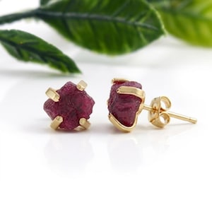 May include: A pair of stud earrings featuring raw, deep red ruby gemstones set in gold metal prongs. One earring is shown from the front, while the other is positioned to display the side profile, including the gold post and butterfly earring back.