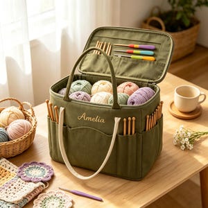 May include: An olive green fabric craft storage bag with the name Amelia embroidered on the front. The bag features multiple exterior and interior pockets filled with wooden crochet hooks, colorful yarn balls, and a purple crochet hook on the table.