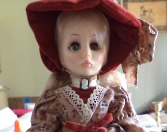 May include: A porcelain doll wearing a red and white dress with a floral pattern. The doll is wearing a red hat with a white ribbon and has blonde hair.