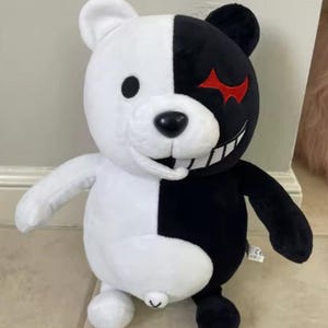 May include: A plush bear toy featuring a split design with one side in white and the other in black. The white side has a round black eye, while the black side features a red jagged eye and a wide, toothy grin with white teeth and a black mouth.
