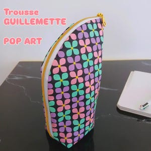 Peut inclure: Une trousse en forme de demi-cercle avec un motif floral répétitif rose, violet et bleu sarcelle sur fond noir. La trousse est dotée d'une fermeture éclair dorée le long du bord incurvé. Le texte "Trousse GUILLEMETTE POP ART" est visible.