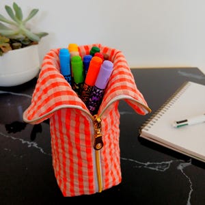 Peut inclure: Une trousse à crayons cylindrique avec un motif vichy orange et blanc et une fermeture éclair dorée. La trousse est ouverte sur le dessus, révélant une collection de marqueurs colorés dans des tons de bleu, vert, orange et violet.